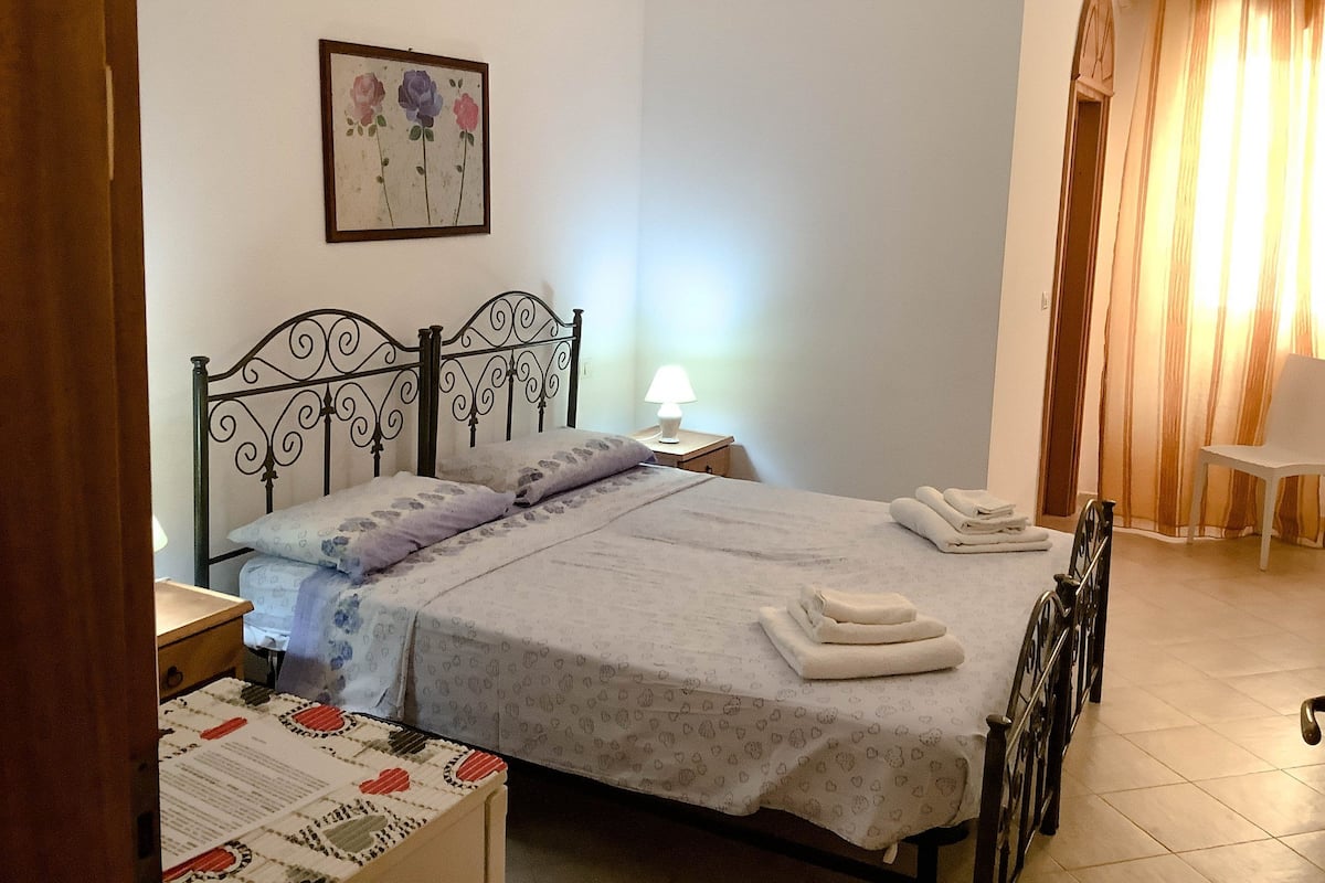 Cama bem arrumada com colcha de padrão floral, abajur e toalha dobrada sobre a cama.