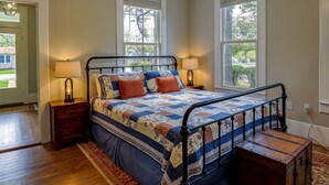 A bedroom with a bed, two bedside tables, a nightstand, a wooden chest, and two windows.