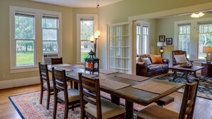 A dining area with a wooden table, chairs, and a rug. There is a sofa and a lamp in the background.