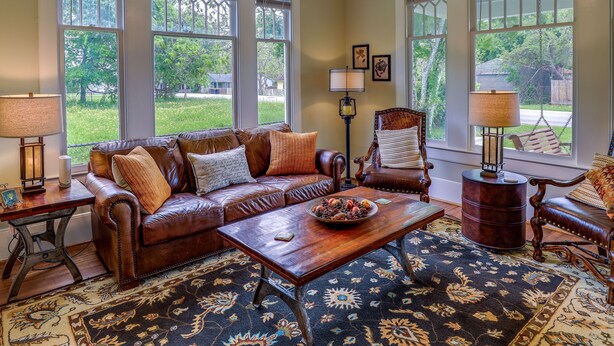A cozy living room with a brown leather sofa, a wooden coffee table, and a patterned rug. There are two windows with white frames, a lamp, and a chair with a cushion.