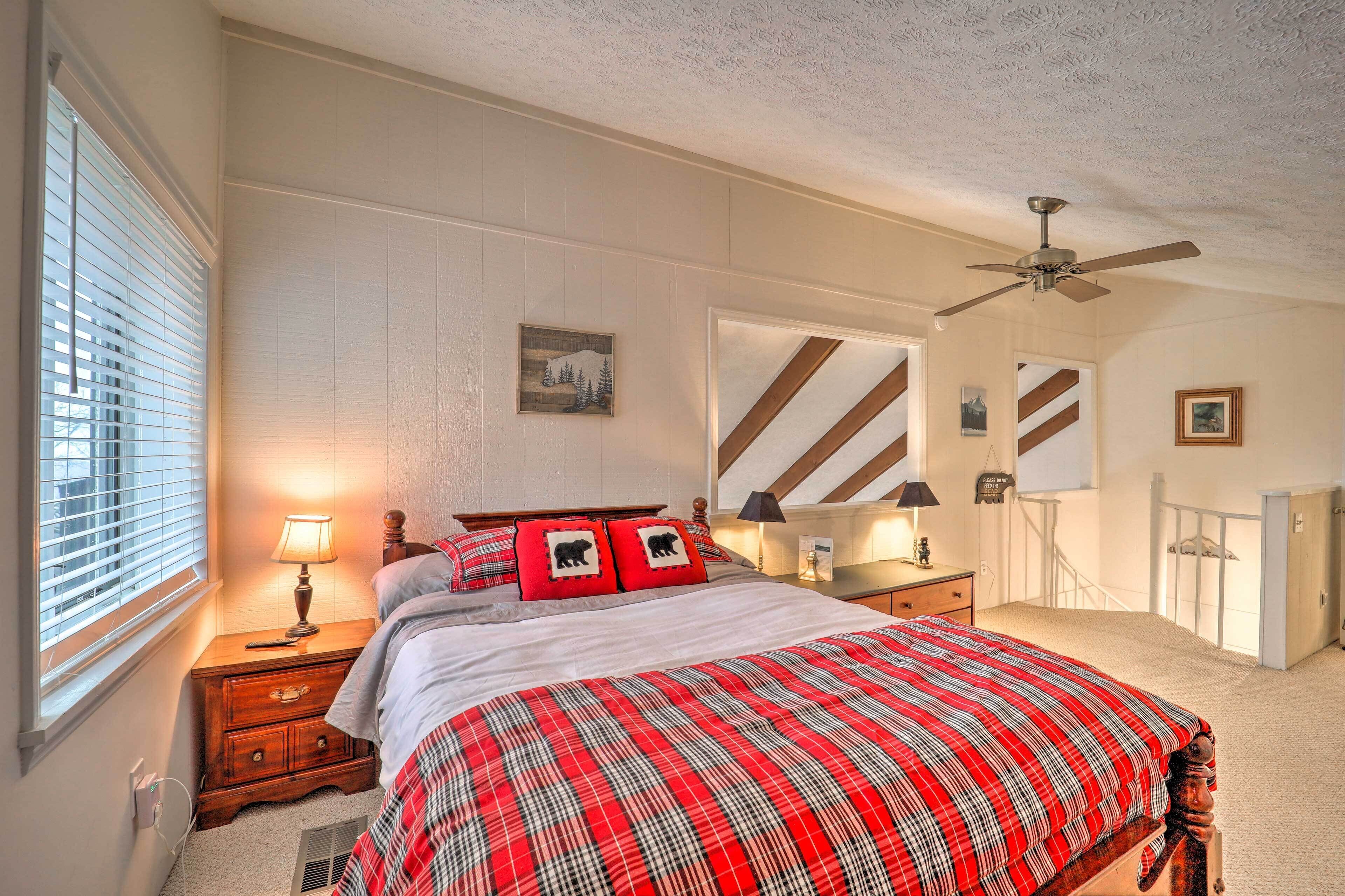 Quarto com cama, mesas de cabeceira, ventoinha no teto, janela com persianas e porta com espelho riscado.