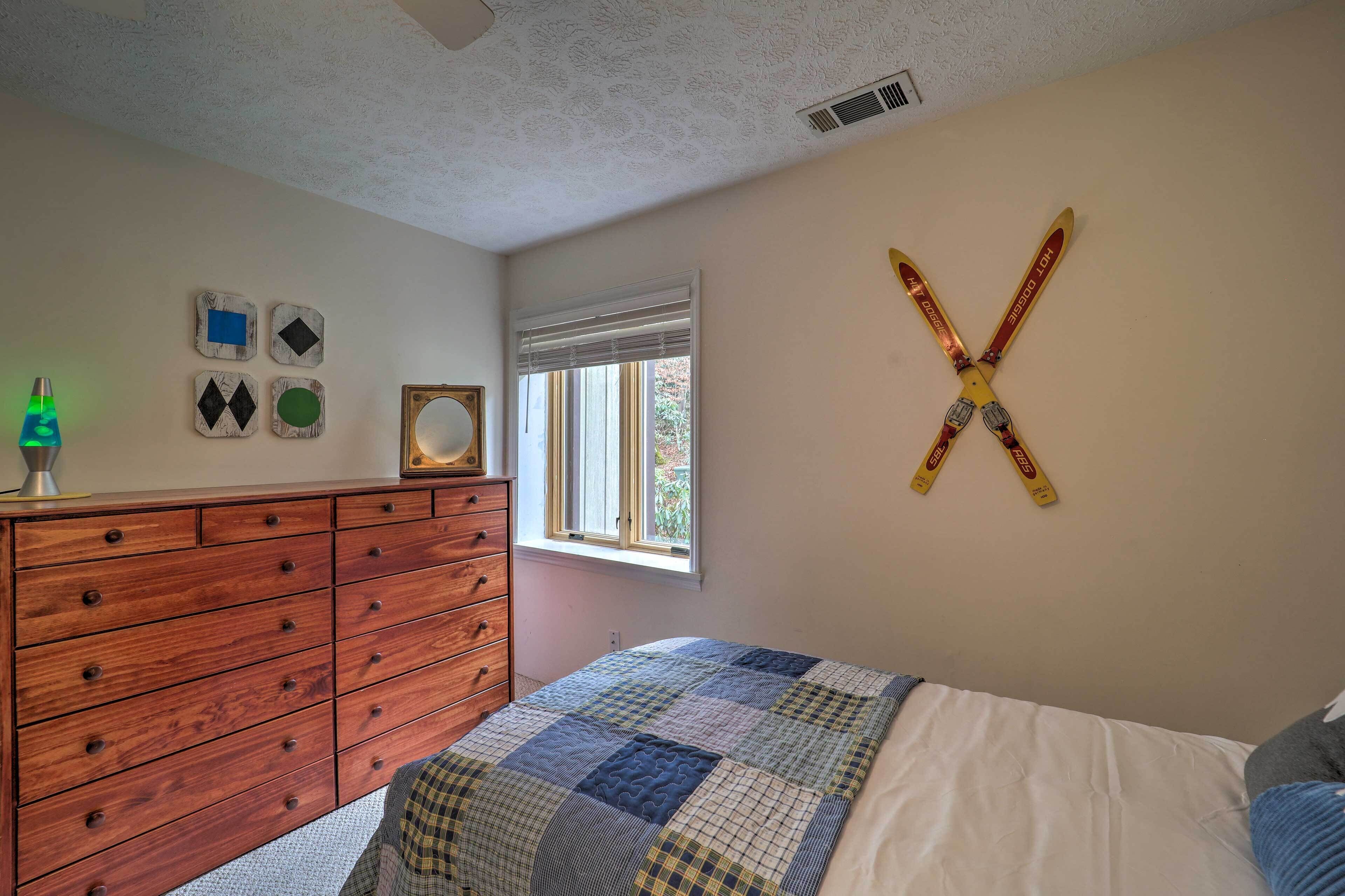 A bedroom with a wooden dresser, a quilted bedspread, a window with blinds, and a wall-mounted ski cross.