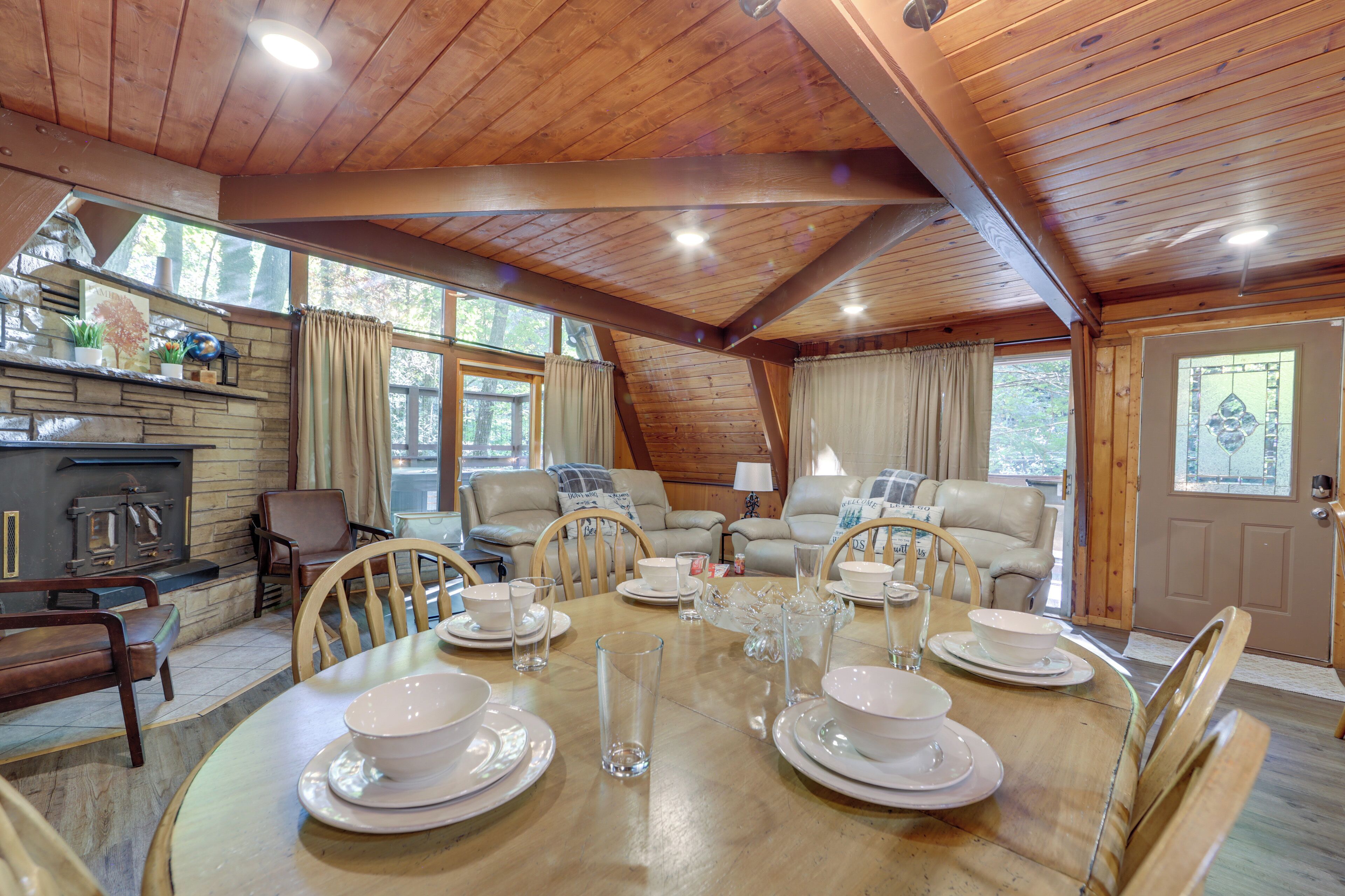 A dining area with a wooden table set for four, surrounded by chairs. There is a fireplace with a stone mantle and a window with curtains.