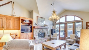 A spacious living room with a stone fireplace, wooden furniture, and large windows offering a view of the landscape. - Seasons Lodge at Arrowhead: Beaver Creek Condo! (Edwards)