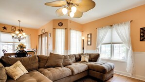 A living room with a sectional sofa, a chandelier, and large windows with curtains. - Breezy Wildwood Condo ~ 2 Blocks to Beach! (Wildwood)
