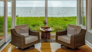 Ein Zimmer mit zwei Korbstühlen, die zu einem großen Fenster mit Blick auf eine Wiese und ein Gewässer zeigen.