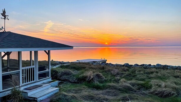 Ein Küstengebäude mit einer überdachten Veranda, Blick auf den Sonnenuntergang über dem Wasser und einer felsigen Küstenlinie.
