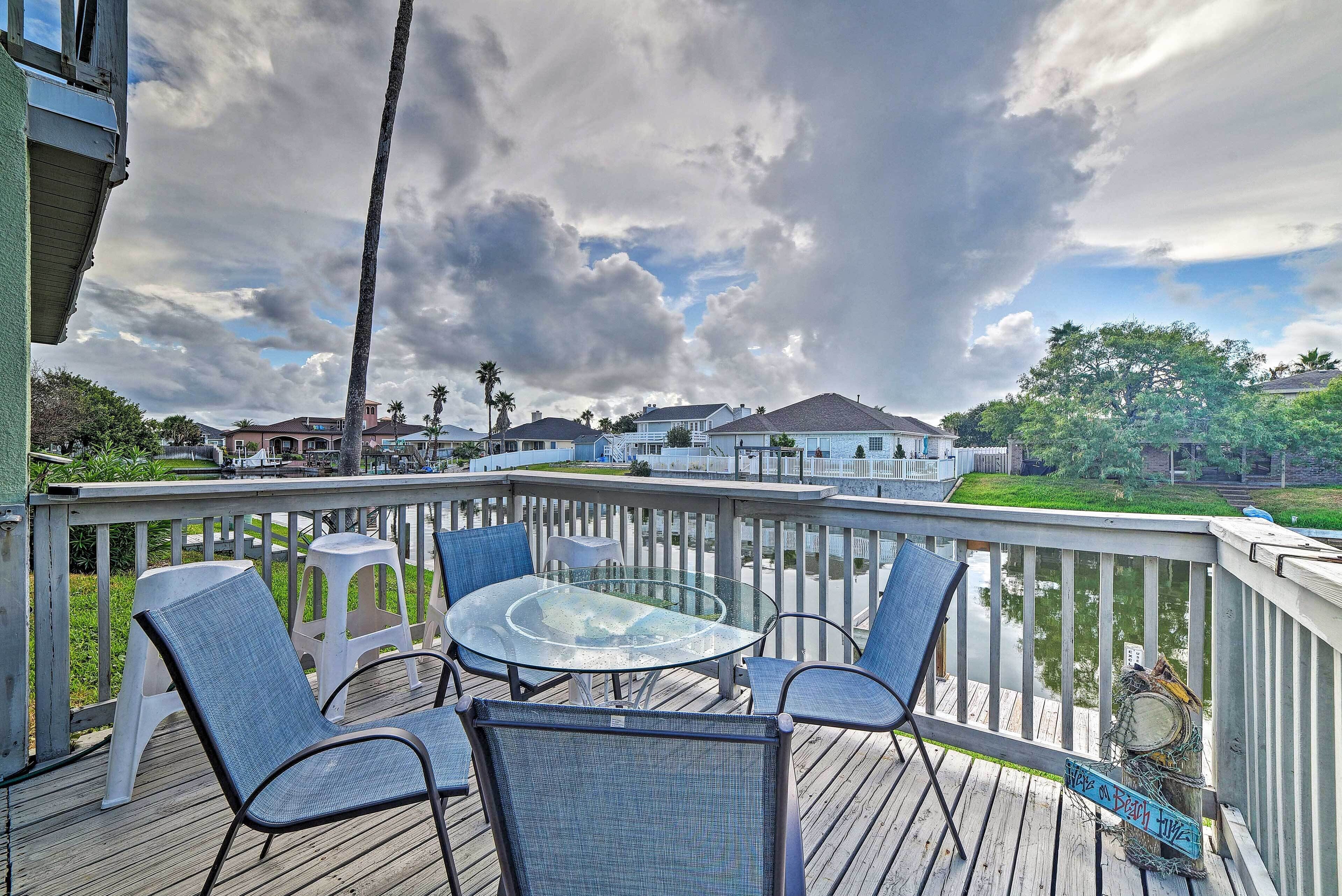 A deck with a glass table and chairs, overlooking a body of water and houses.