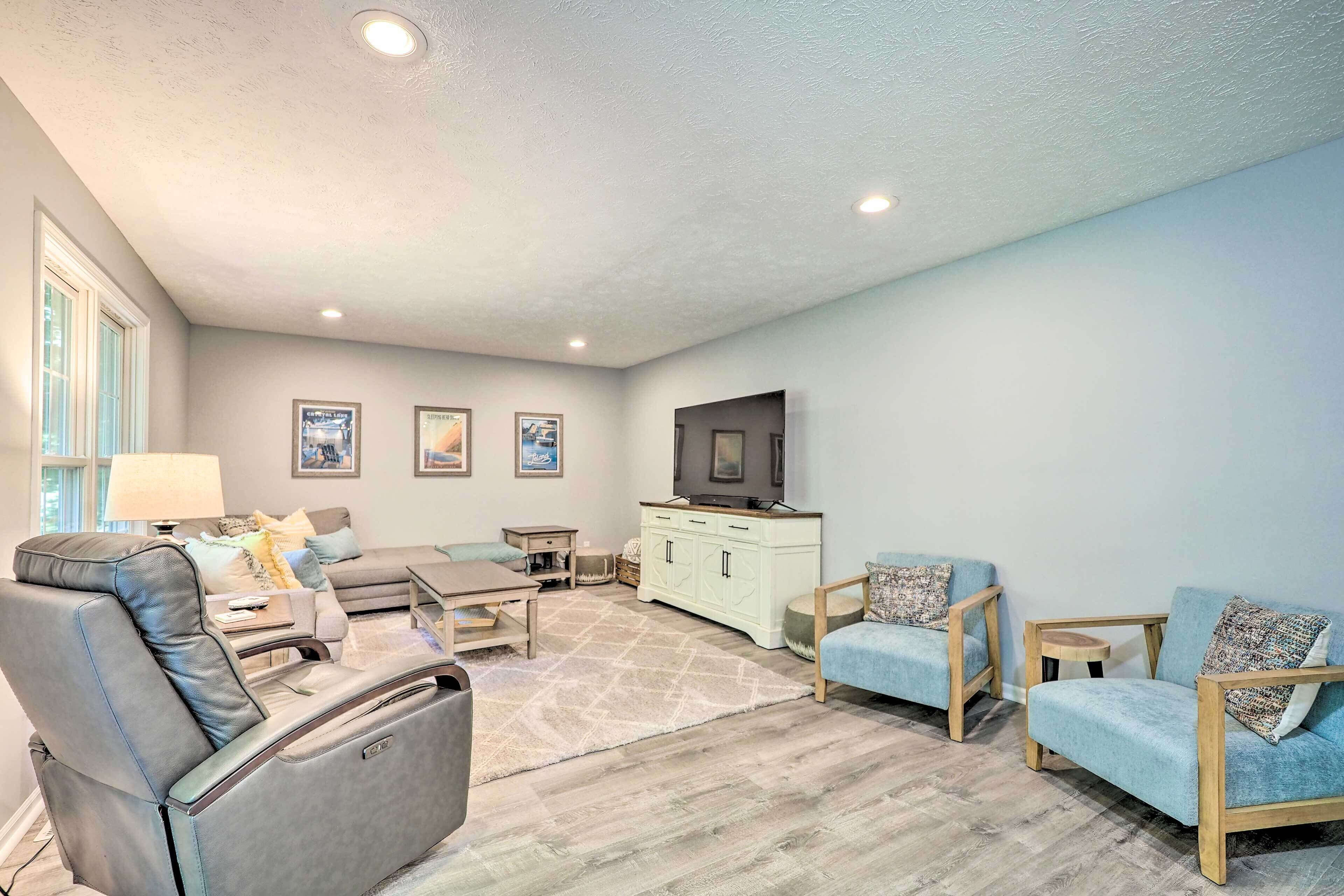 A spacious living room with a leather recliner, a sofa, and two armchairs. A television is mounted on the wall, and there are framed pictures on the wall.