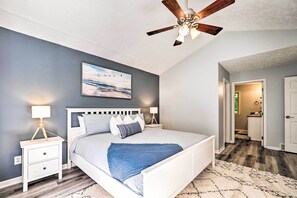 Une chambre à coucher comprenant un lit, des tables de chevet, un ventilateur de plafond, un tableau et un couloir menant à une salle de bain.