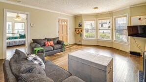 Un salon spacieux avec un grand canapé d’angle gris, un petit tabouret vert et une commode de rangement grise. Plusieurs fenêtres sont équipées de stores, et le salon comprend également un ventilateur de plafond et un téléviseur à écran plat fixé au mur.