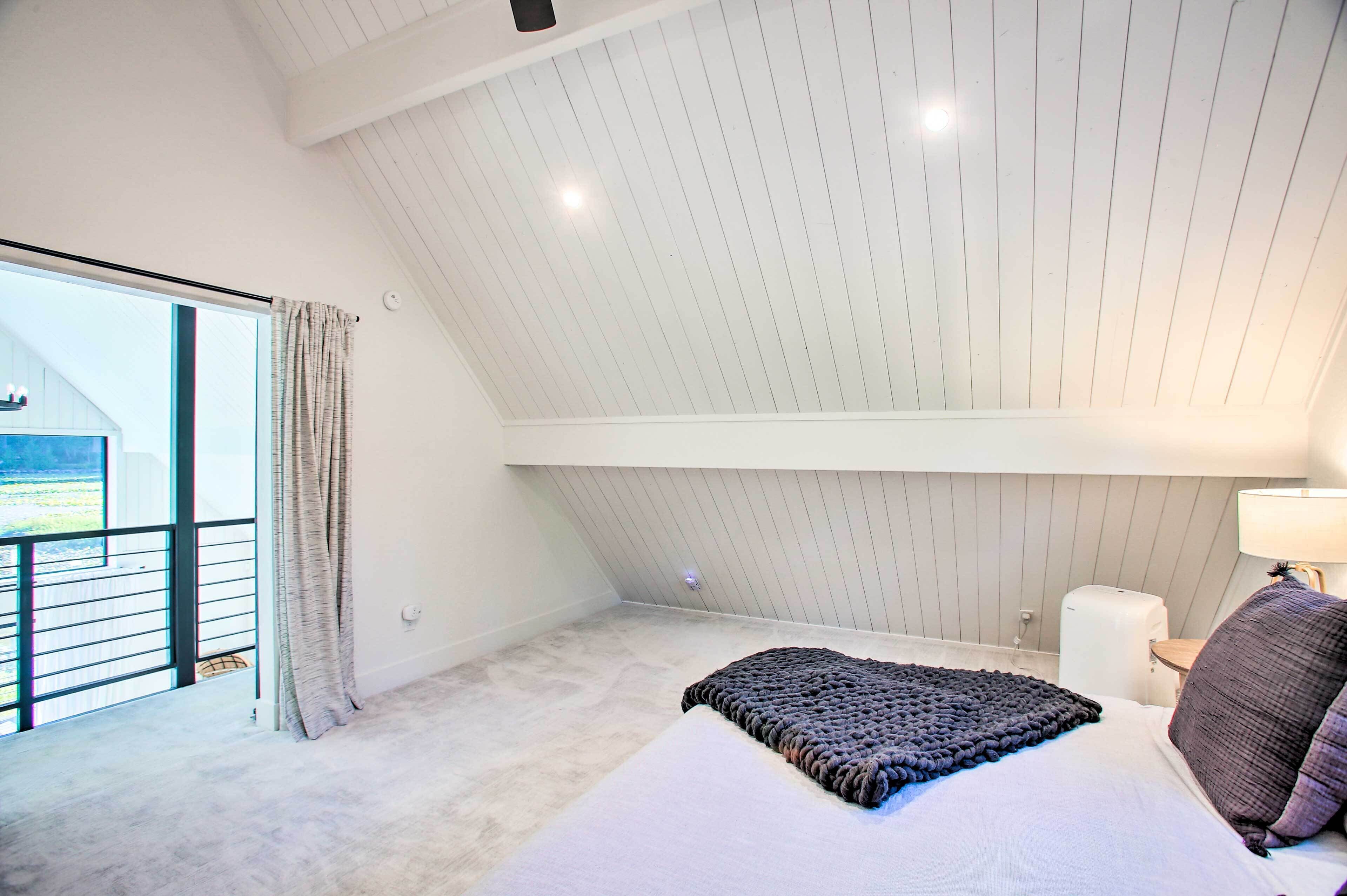 Quarto com teto inclinado, uma janela grande, uma cama com cobertor, mesa de cabeceira e abajur.