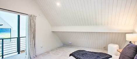 Quarto com teto inclinado, uma janela grande, uma cama com cobertor, mesa de cabeceira e abajur.