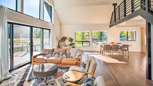 A spacious living area with a large sofa, a round coffee table, and a dining set. There are multiple windows providing natural light and views of the outdoors. - A Contemporary Dream: Lakefront Rathdrum Oasis! (Rathdrum)