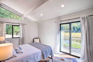 A bedroom with a bed, a nightstand, a lamp, and a view of a garden through large windows. - A Contemporary Dream: Lakefront Rathdrum Oasis! (Rathdrum)