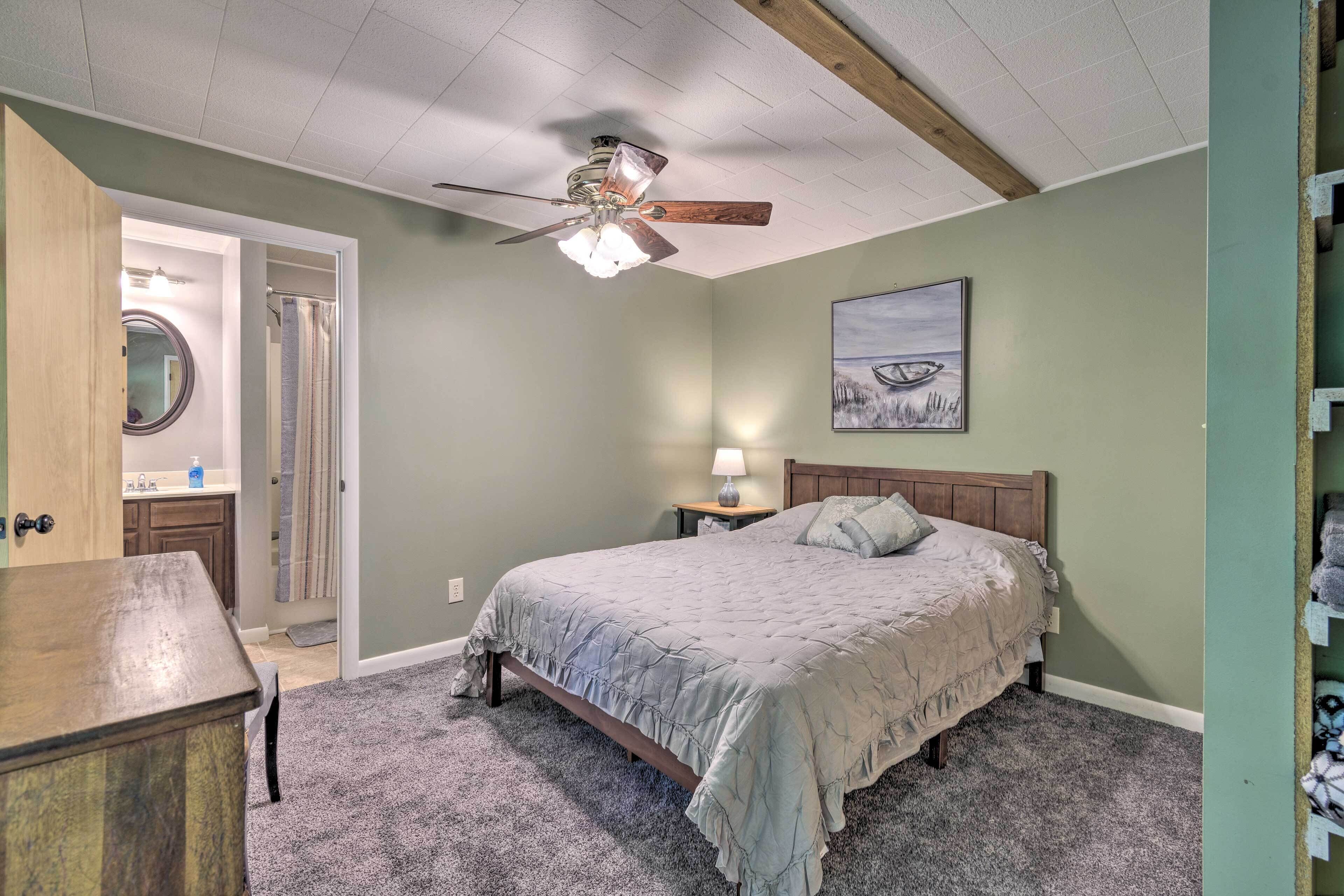 A bedroom with a bed, a ceiling fan, a nightstand with a lamp, a wooden dresser, a bathroom with a mirror, and a painting on the wall.