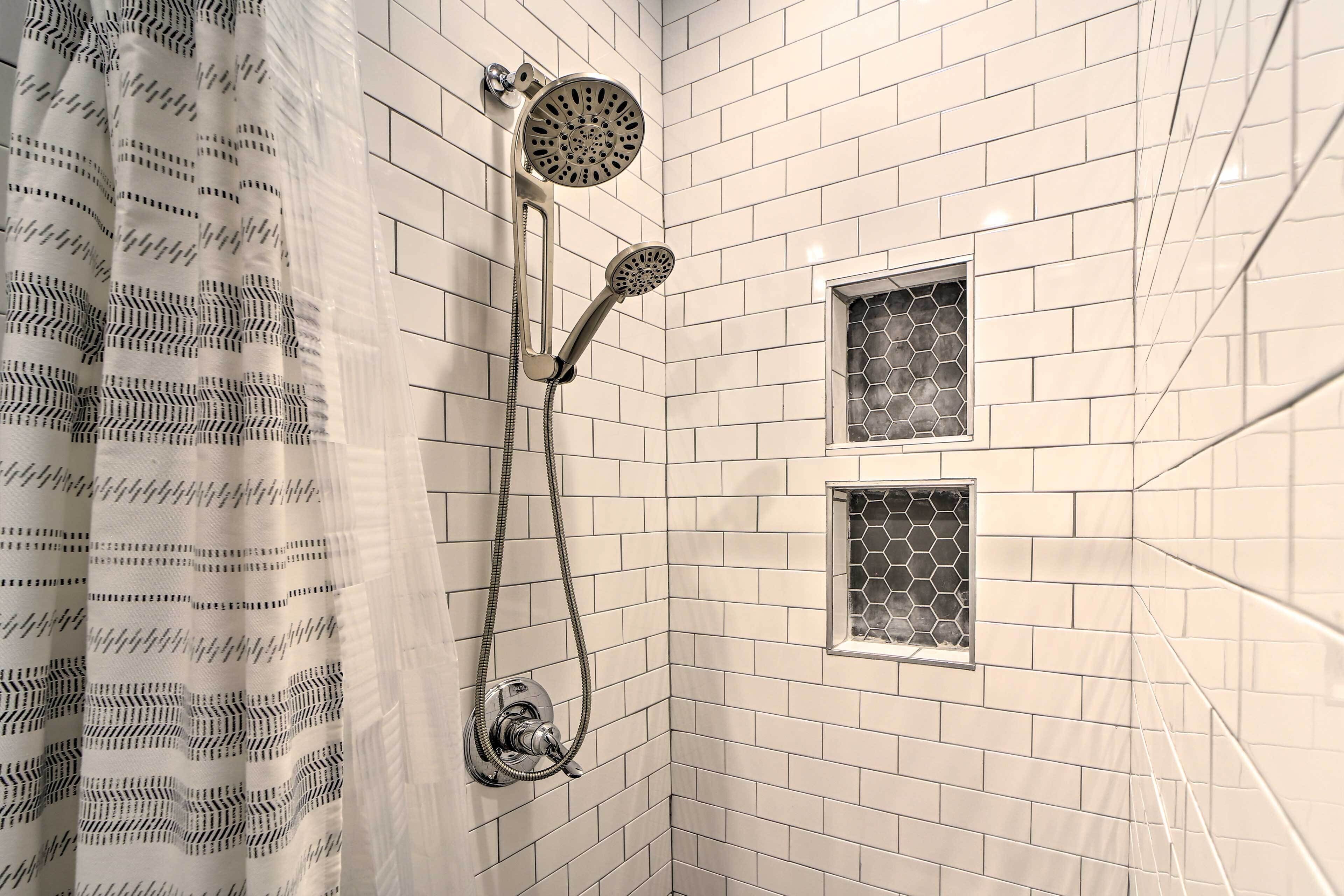 A bathroom with white tiled walls, a shower with a handheld showerhead, and a window with a hexagonal pattern.
