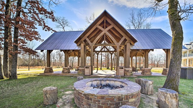A pavilion with a brick fire pit in the center, surrounded by wooden posts, set in a grassy area with trees and a building in the background. - Cozy Carlton Landing Home - Near Lake Eufaula (Eufaula)