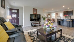 A modern living room with a dark brown sofa, a coffee table with a vase of flowers, a flat-screen TV, and a dining area with a bar. - 9 Mi to Duke University: Spacious Home w/ Yard! (Durham)