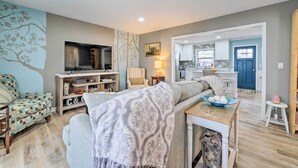A living room with a sofa, a television, and a dining table with a bowl. There is a kitchen in the background with cabinets and a blue door. - Cozy Wilmington Vacation Rental: 10 Mi to Beach! (Wilmington)