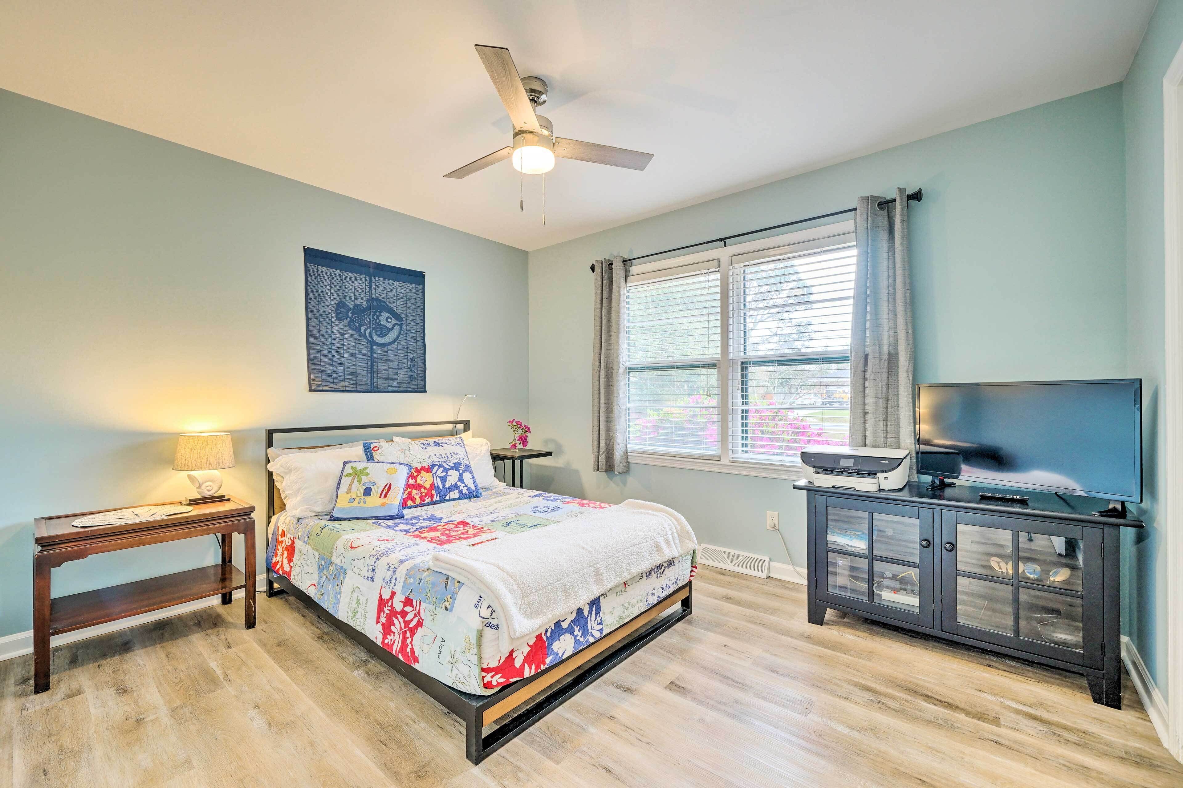 A bedroom with a bed, a nightstand, a TV stand, a ceiling fan, and a window with curtains.
