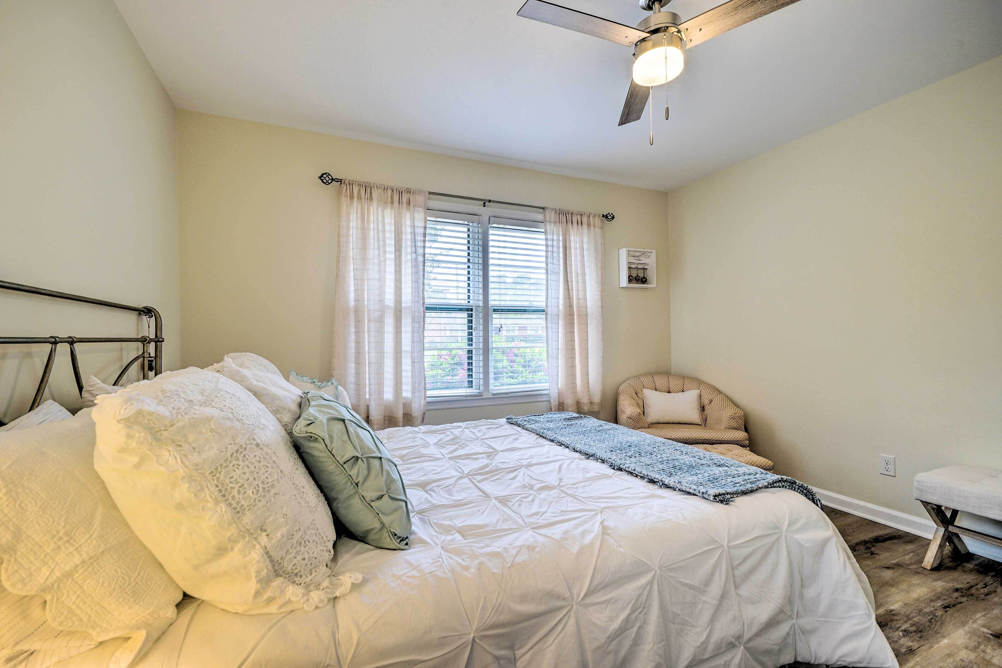 Ein Schlafzimmer mit Bett, Stuhl, einem Vorhangfenster, Deckenventilator und Wandleuchte.