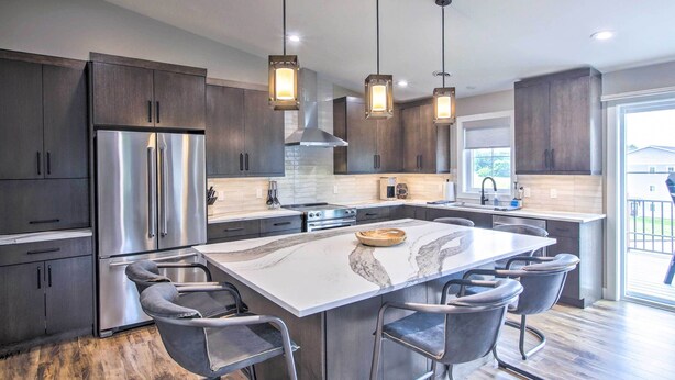 A modern kitchen with dark wood cabinets, stainless steel appliances, a marble island with bar stools, pendant lighting, and a large window providing natural light. - Chic Plymouth Townhome < 1 Mi to Road America (Plymouth)