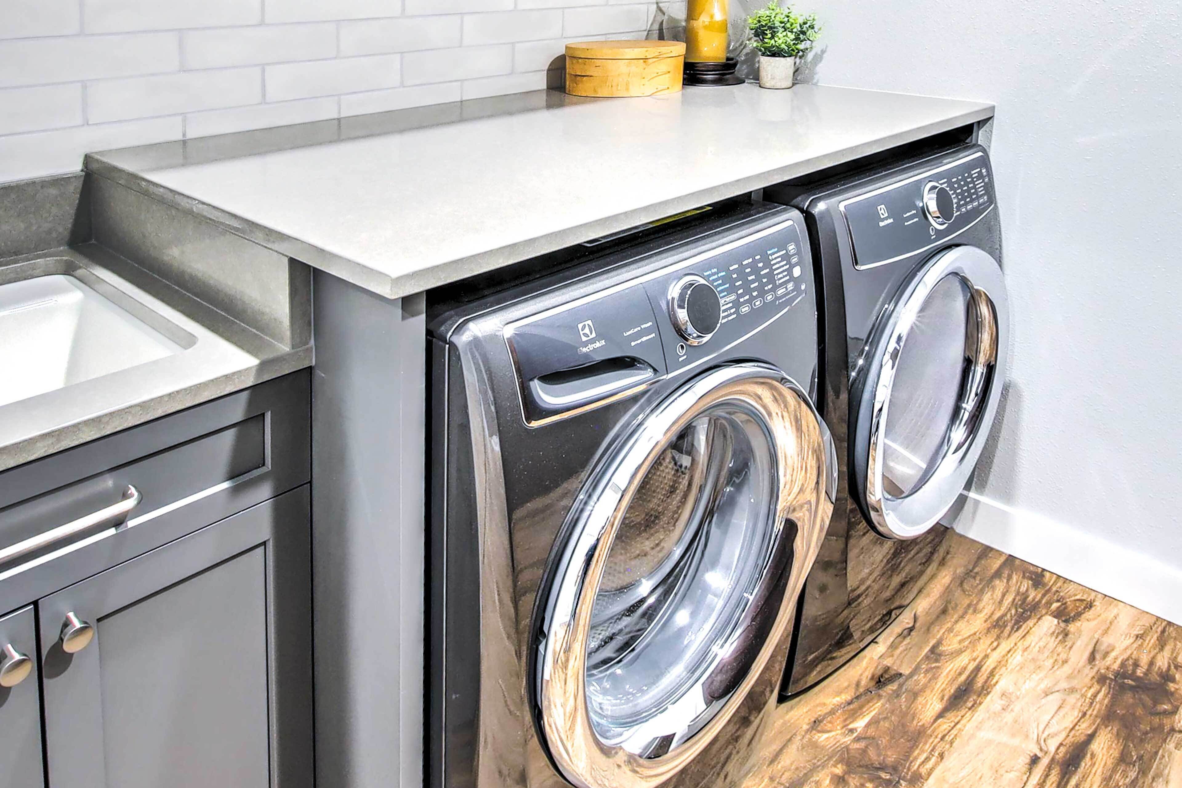 A compact washer and dryer unit with a stainless steel finish, integrated into a kitchen counter with a sink and cabinets.