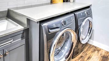 A compact washer and dryer unit with a stainless steel finish, integrated into a kitchen counter with a sink and cabinets.