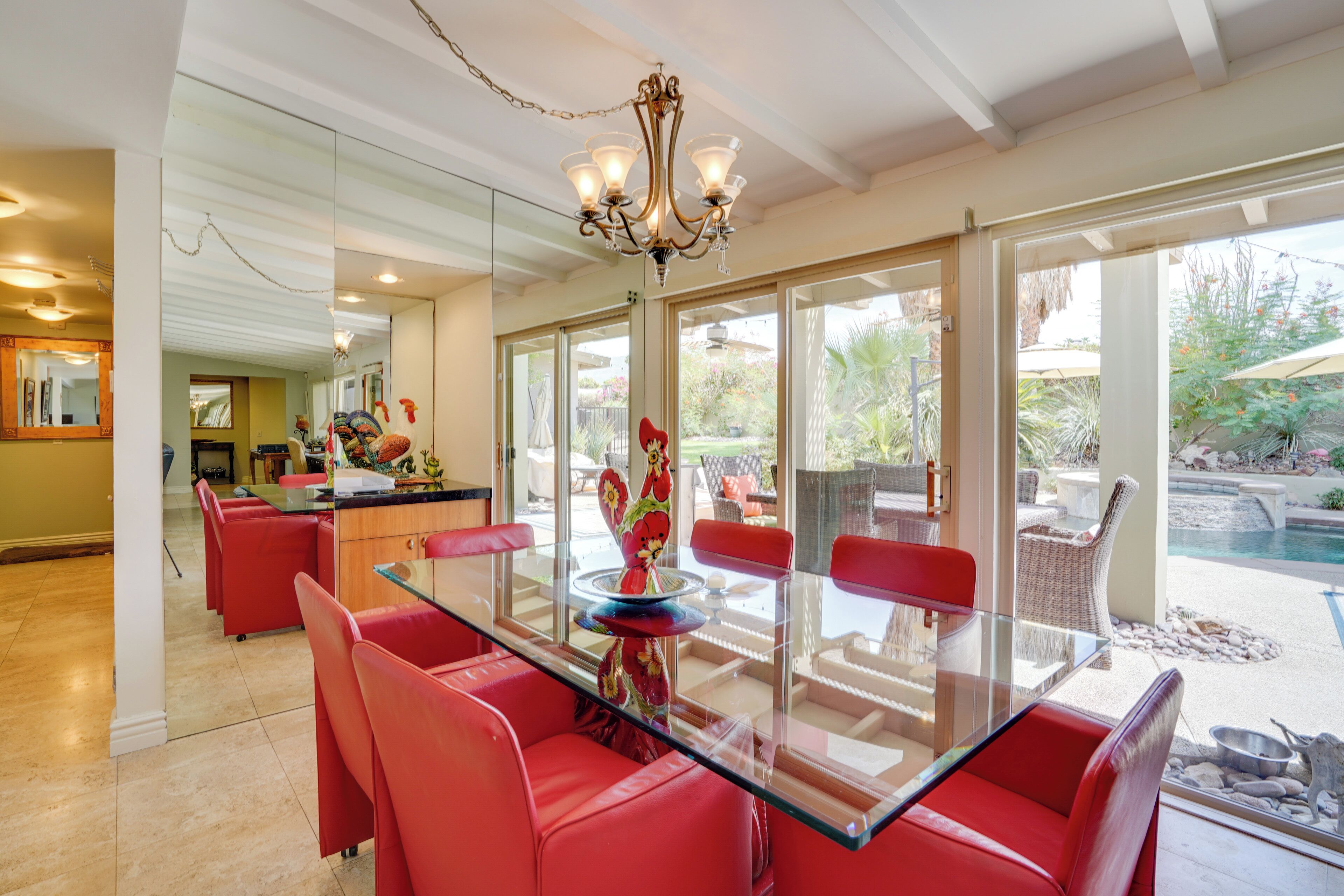 A dining area with a glass table and red chairs, a chandelier, and a view of the outdoor patio.