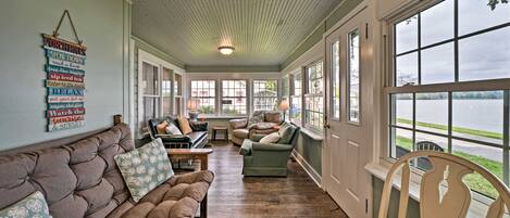 A cozy living room with a large window, a cushioned sofa, a wooden chair, and a wall-mounted sign with various phrases.