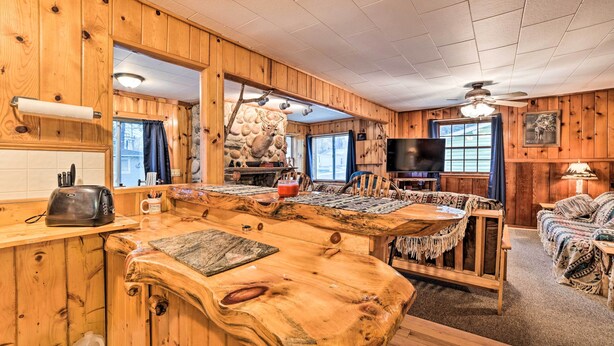 A rustic cabin interior with a wooden dining table, a stone fireplace, a television, and a cozy seating area.