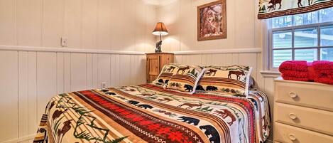 A bedroom with a large bed, a nightstand, a lamp, a window with a view, and a wall with a painting.