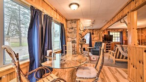 A wooden cabin interior with a glass table, rustic chairs, a stone wall, and a view of the outdoors.