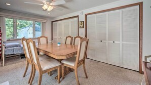 Comedor con una mesa y sillas de madera, un ventilador de techo y vista a una casa a través de amplios ventanales.