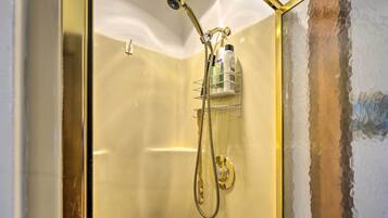 A shower with a gold showerhead and hose, a glass door, and a shelf with toiletries.
