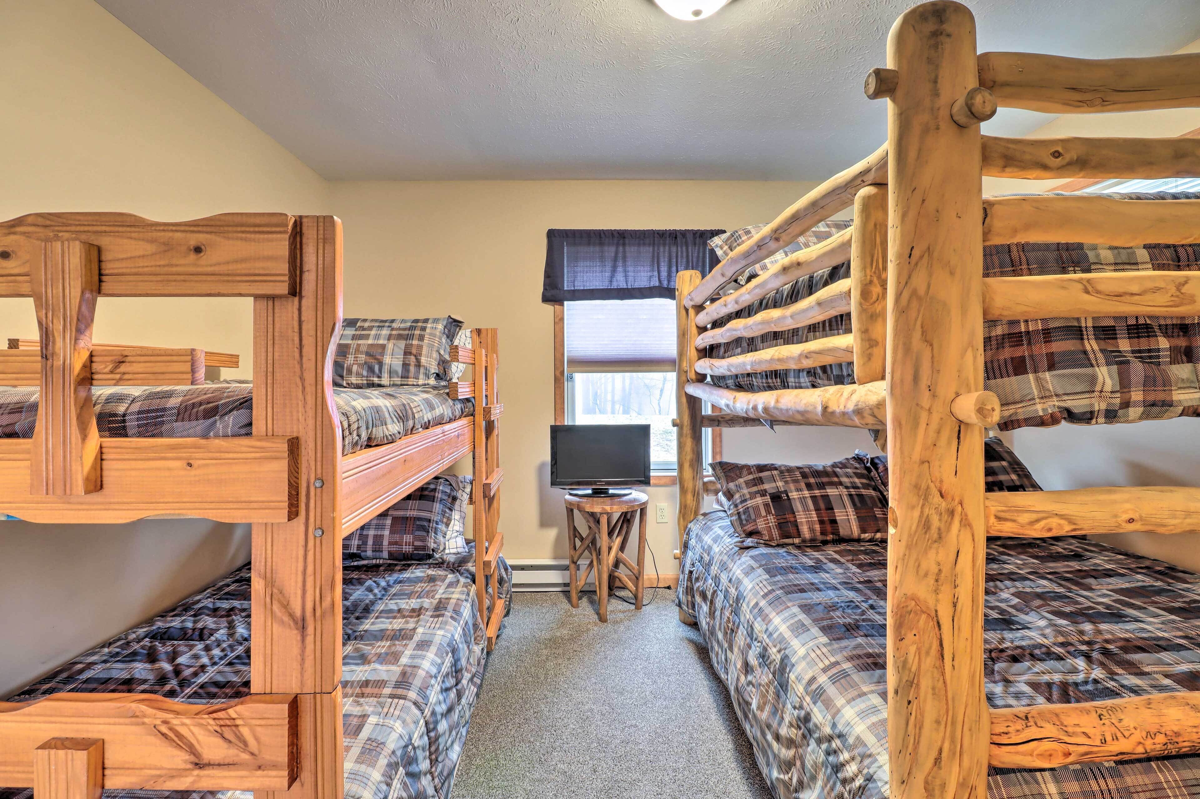 A room with three wooden bunk beds, a small wooden table with a television, and a window with a view of trees.