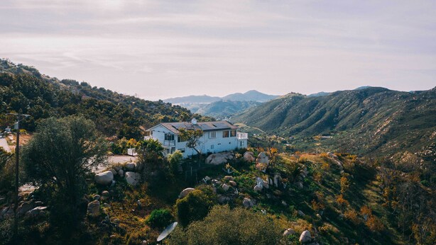 A white house with a sloped roof, surrounded by rocky terrain and greenery, with mountains in the background.