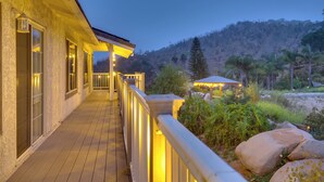 A wooden deck with a railing, a building with large windows, and a view of a mountainous landscape with trees.