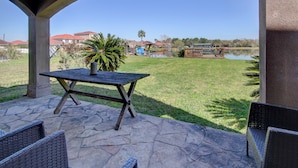 A patio with a stone table and wicker chairs, overlooking a grassy area with a lake and houses in the background.