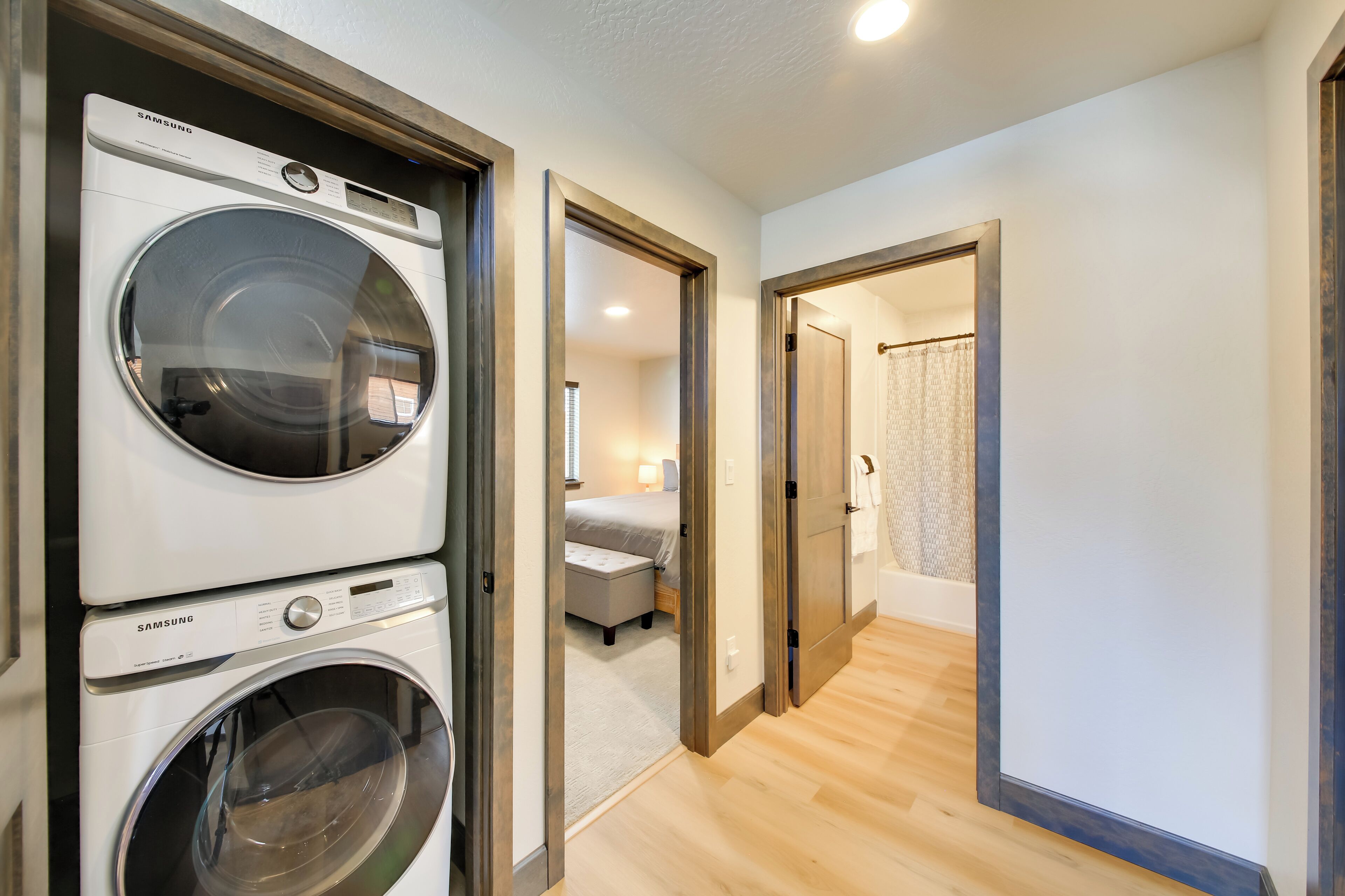 A laundry area with two Samsung washing machines, a hallway leading to a bedroom with a bed and nightstand, and a bathroom with a shower curtain.