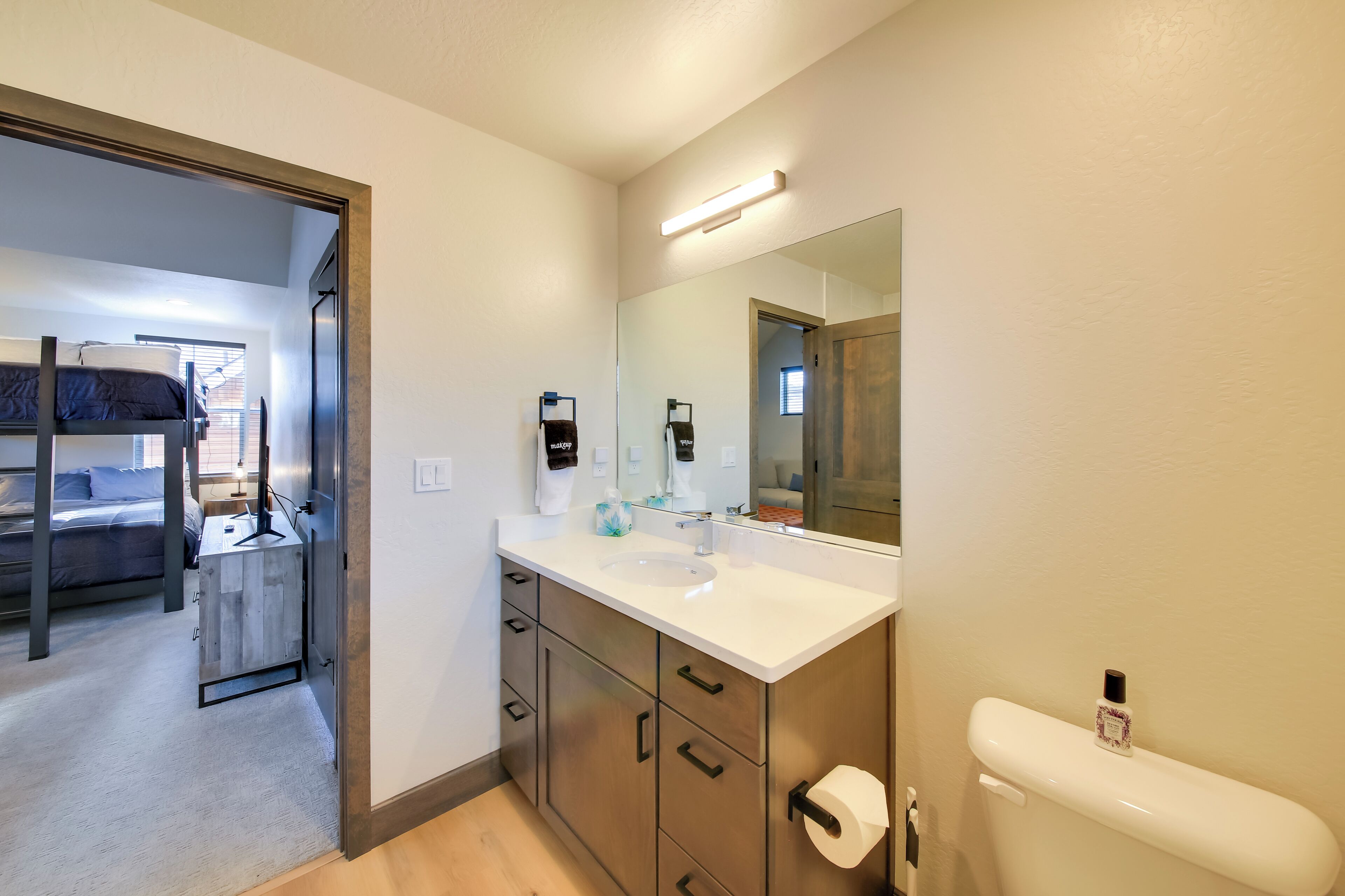A modern bathroom with a large mirror, a sink, and a toilet. There is a wooden cabinet with drawers and a small shelf.
