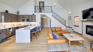 Ein großzügiger Wohnbereich mit einer großen Couch, einem Couchtisch und einem Kamin. Es gibt eine Küche mit modernen Geräten und einen Essbereich mit einer großen Kochinsel. Eine Treppe führt auf eine obere Ebene.