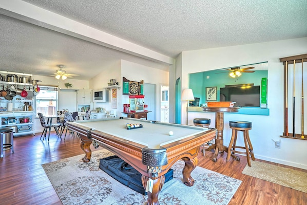 A spacious living area with a pool table, wooden flooring, and a kitchen in the background.