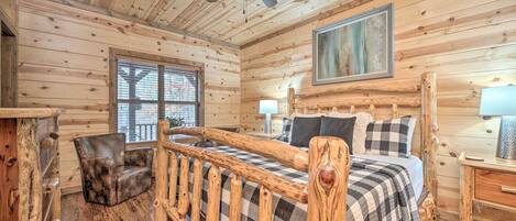 A wooden cabin bedroom with a bed, a chair, a ceiling fan, a window, and a painting on the wall.