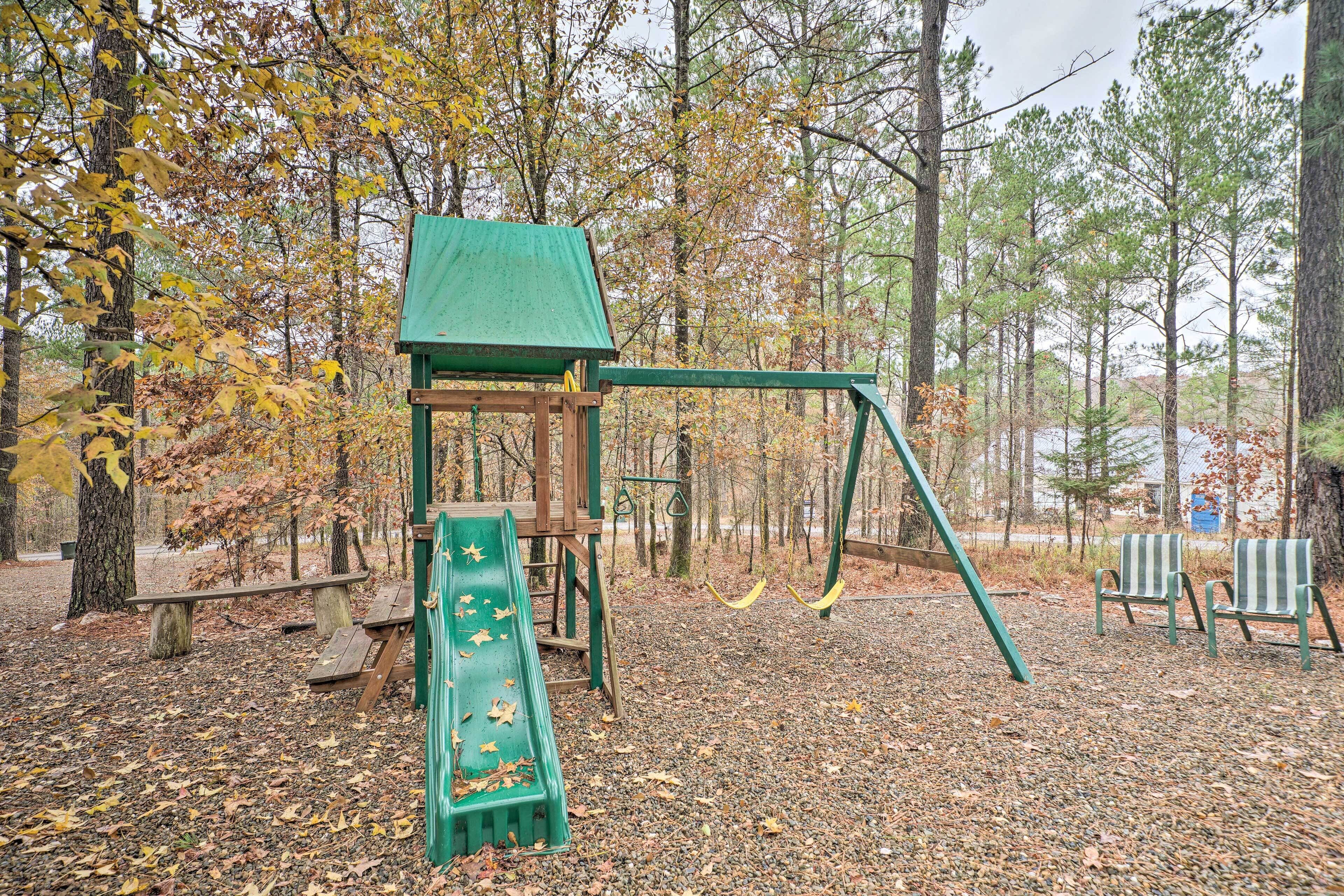 A playground with a slide, swings, and benches in a wooded area.