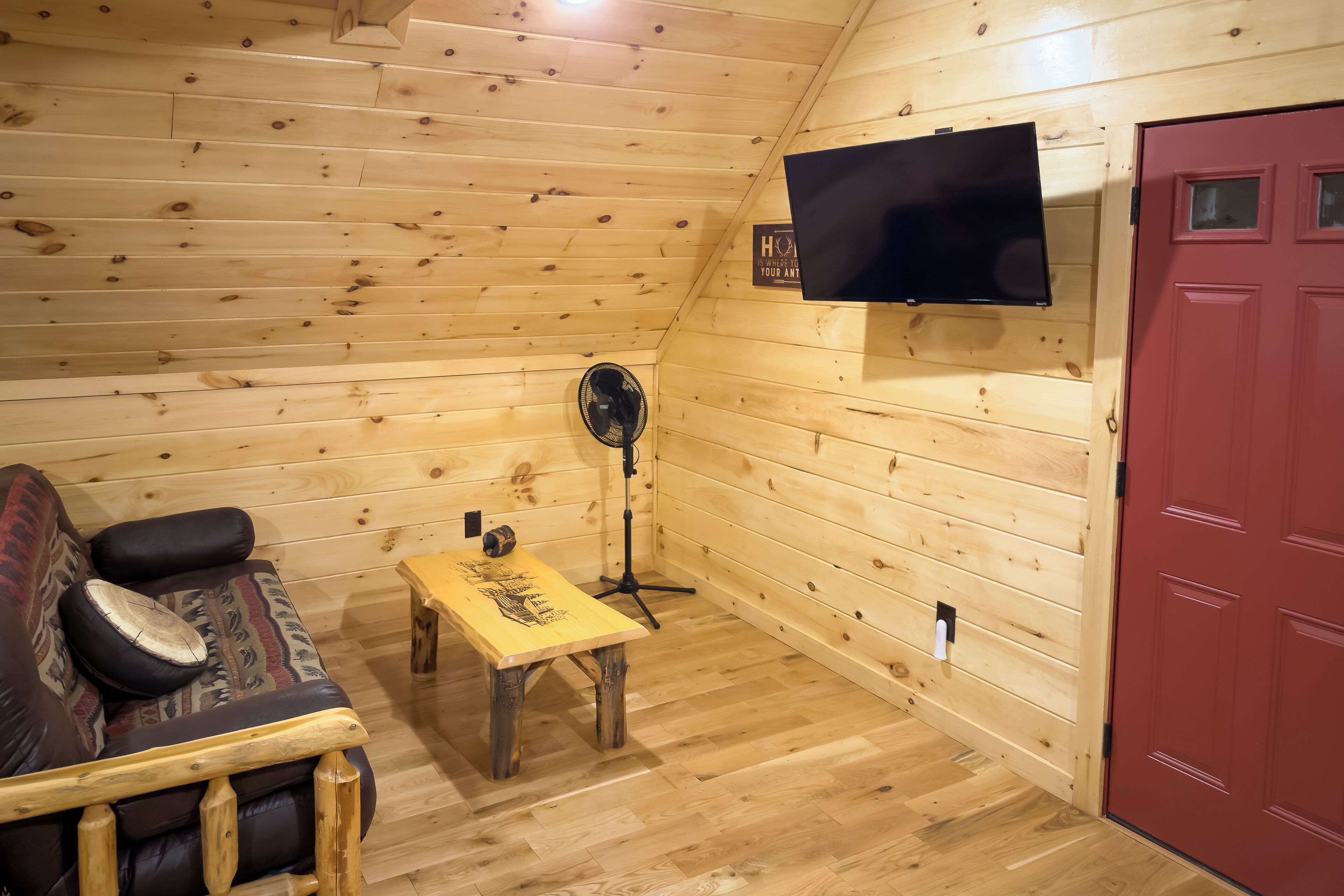 A wooden cabin interior with a sofa, a small table, a fan, and a TV mounted on the wall.