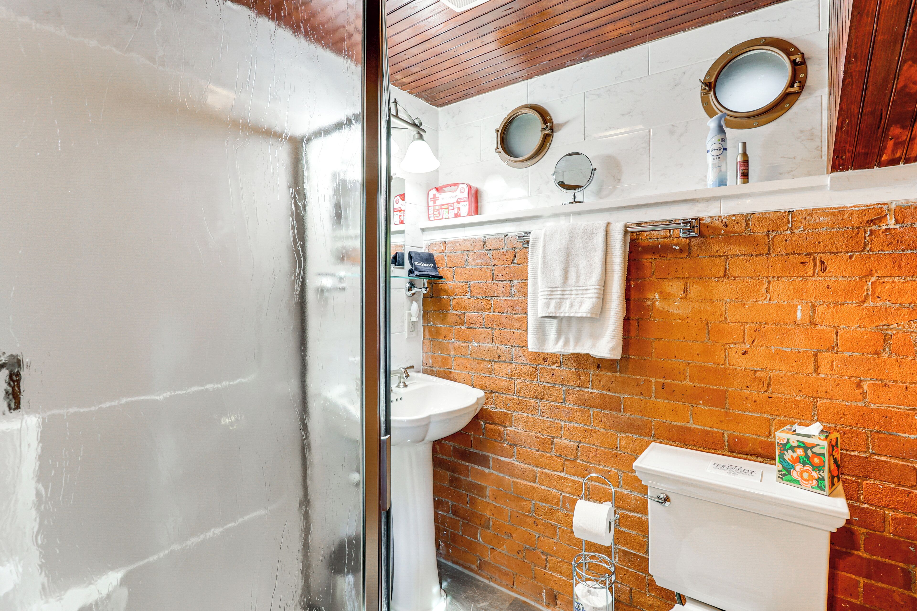 Ein Badezimmer mit einem weißen Waschbecken, einer Toilette und einer Ziegelwand. Zwei runde Spiegel sind an der Wand angebracht, und ein Handtuch hängt dort.