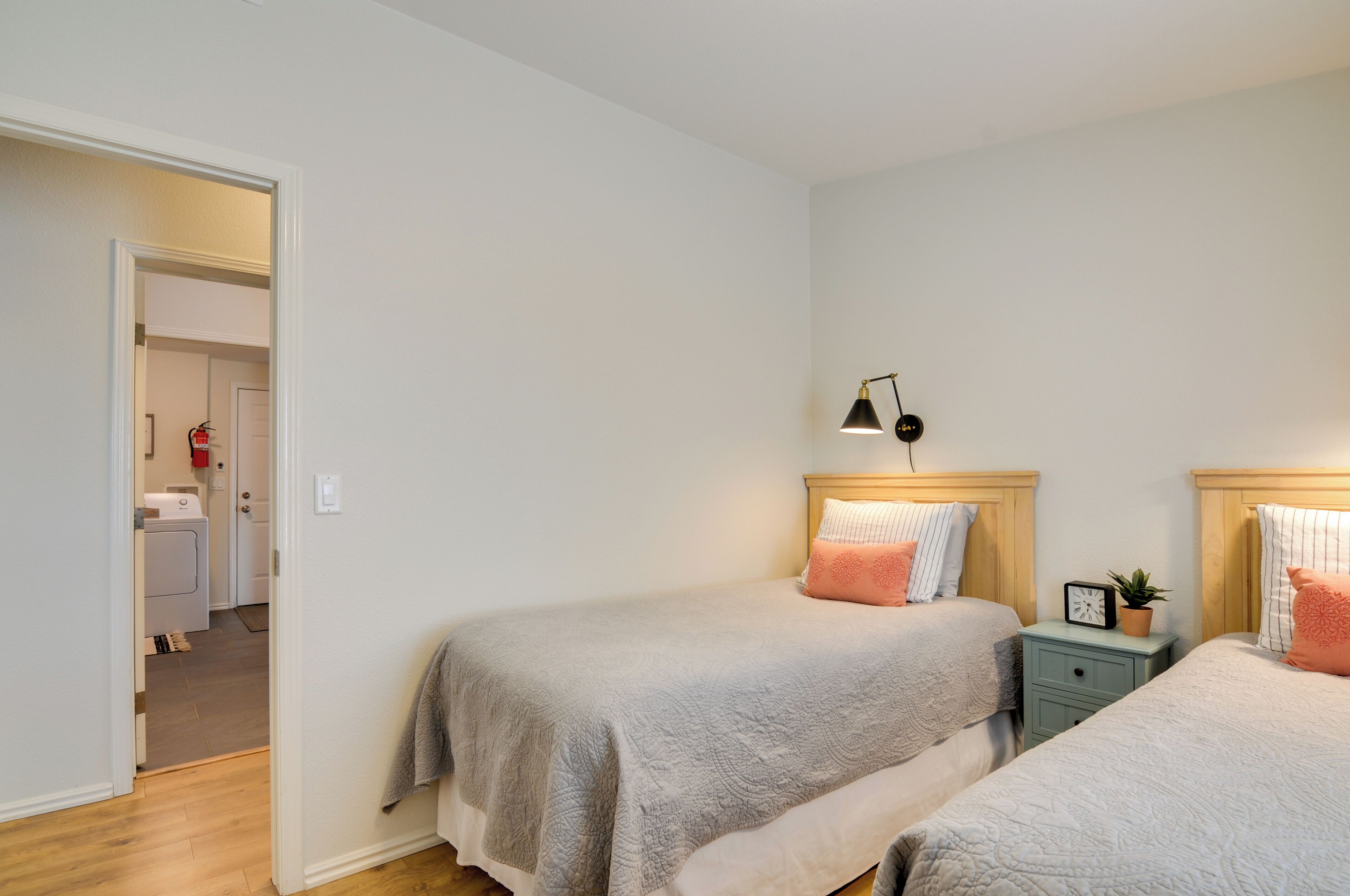 Een slaapkamer met twee bedden, een nachtkastje met een klok en een plant, en een deur naar een wasruimte.