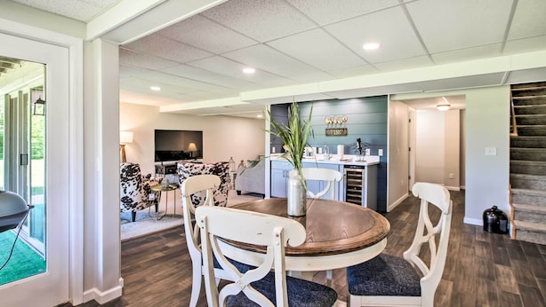 A dining area with a round table and white chairs, a kitchen in the background, and a staircase leading to another level.
